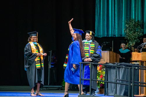 A Clark graduate strides confidently onto the stage, waving to the audience with a bright smile as she nears Dr. Edwards to receive a scroll.