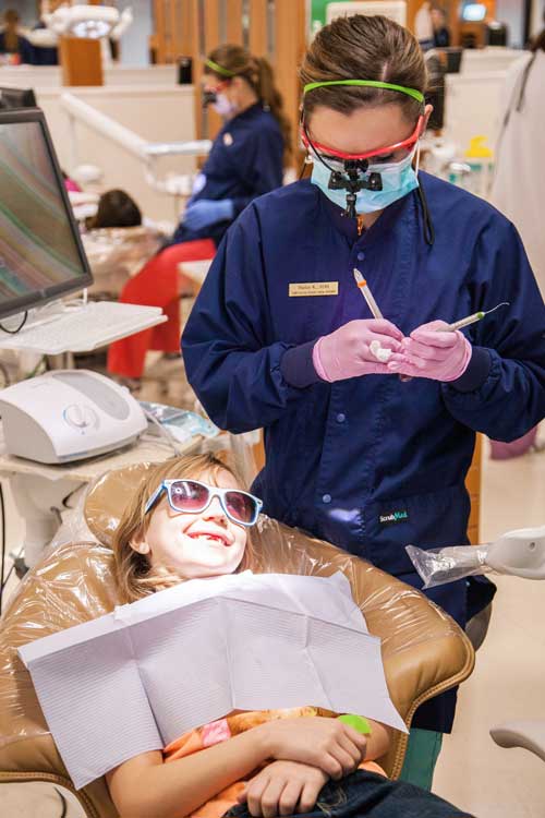 A smiling child wearing protective eyewear sitting in a dental chair while a Clark student hygienist cleans their teeth.